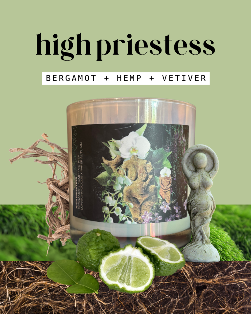 the high priestess candle is pictured in white, iridescent glass featuring a label with a dynamic green collage of two ancient-looking stone figures of voluptuous women's bodies. the candle is surrounded by green moss, bergamot fruit, vetiver roots, and stone. the scent notes say "bergamot + hemp + vetiver"